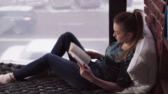 Young Pensive Woman Reading Book on Bed By Window Overlooking City Street