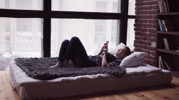 Sad Woman Using Smartphone, Lying on Bed By Window, Overlooking City Street
