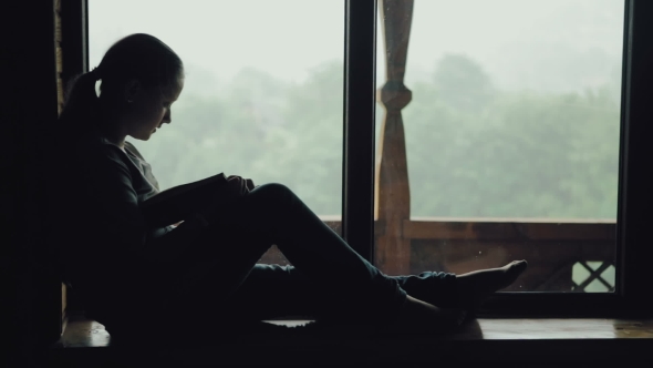 Cute Girl Reading a Book on a Wooden Window Sill on a Rainy Day.