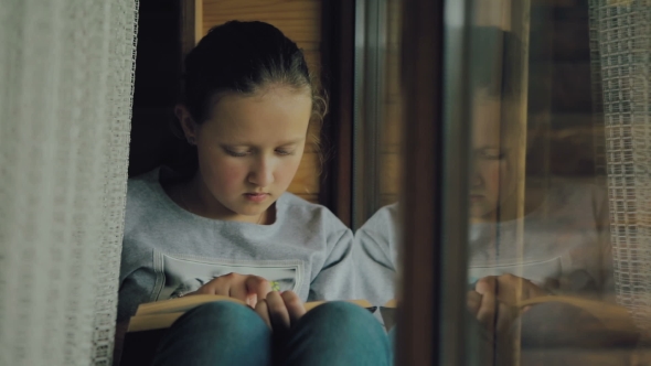Sweet Girl with Blue Eyes Reads a Book on the Window Sill. The Girl Reads a Book and Then Looks Into