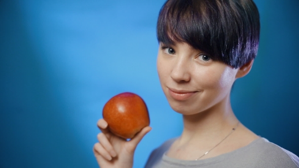Attractive Brunette Girl Holding a Red Apple and Laughing on a Blue Wall