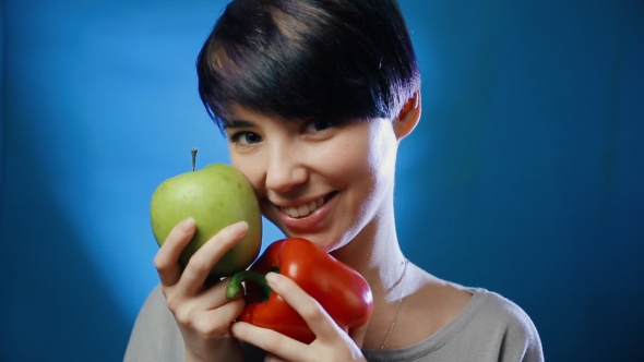 Attractive Brunette Girl Holding a Green Apple and Red Pepper Laughing on a Blue Wall