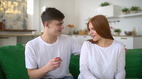 Young Man Making Marriage Proposal To Surprised Woman with Ring in Red Gift Box Sitting on Sofa alt
