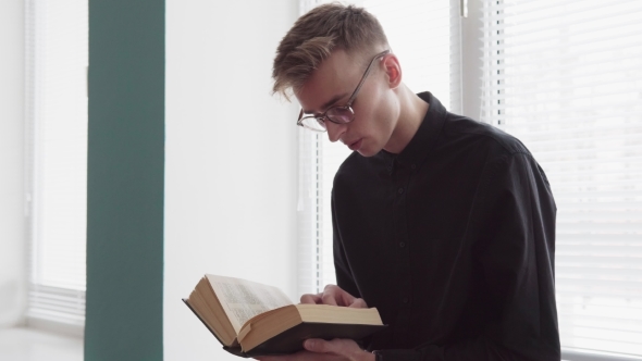 Young Serious Man Is Reading a Bible Against a Window