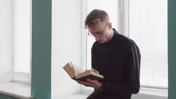 Young Handsome Man Is Reading a Book Against a Window