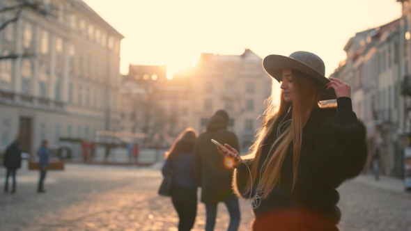 Young Woman in a Bright Sunlight Uses Her Phone While in the City Center alt