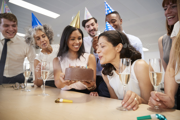 Woman blows out candles on birthday cake in office Stock Photo by monkeybusiness