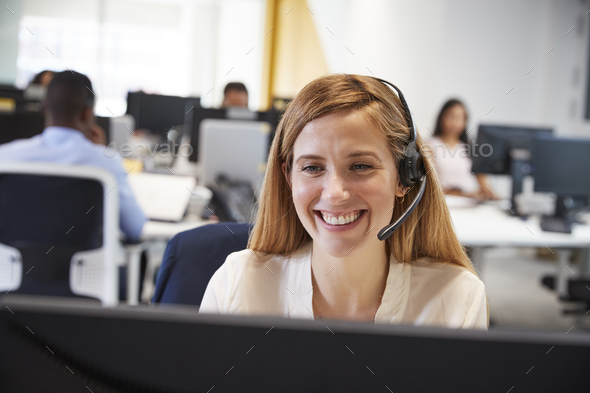 Young woman working at computer with headset in busy office Stock Photo by monkeybusiness
