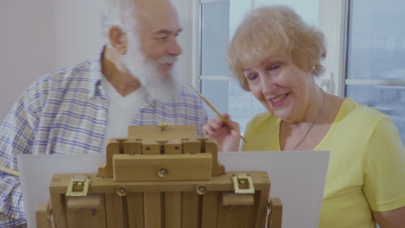 Senior Man Has Fun with His Wife Drawing the Picture on Easel alt