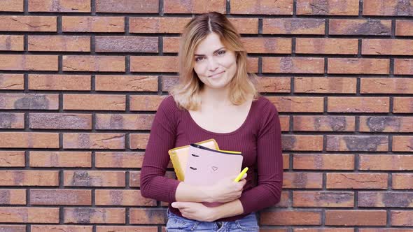 Woman Stands Holding Notebooks and Pen Near Brick Wall