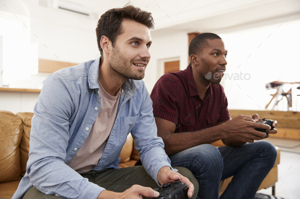 Two Male Friends Sitting On Sofa In Lounge Playing Video Game Stock Photo by monkeybusiness