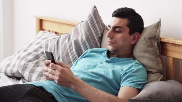 Young Middle Eastern Male Using a Smartphone, Lying on Bed at Home
