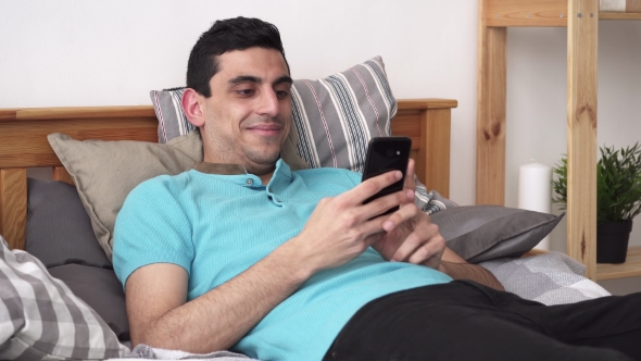 Happy Arabic Man Using a Smartphone, Lying on Bed at Home