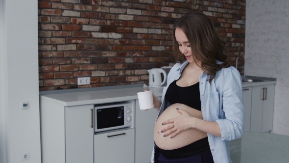 A Beautiful Pregnant Woman with an Open Stomach Is Standing in Her Kitchen at Home and Drinking alt
