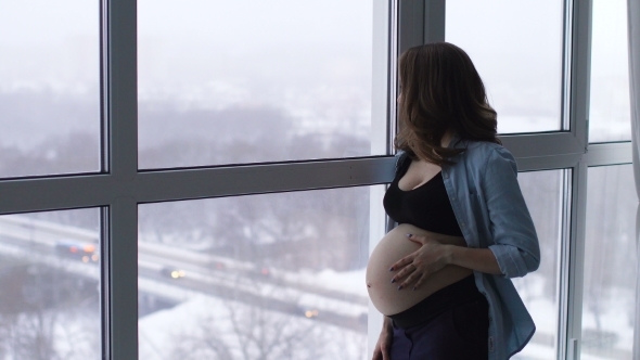 A Happy Pregnant Woman Stands at a Large Panoramic Window Located at a High Altitude. He Touches His alt