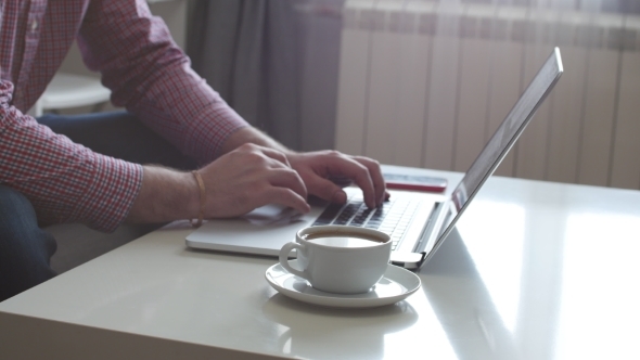A Person Works From Home Using the Top and Trackpad. Young Business Man Sitting at Table Drinking alt