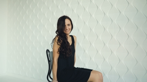 Brunette Young Women Poses, Smiling, Waving Hair, Sitting and Turning on the Chair in Black Dress