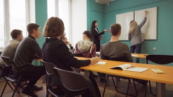 Woman Student Writes on a Whiteboard in Front of a Teacher and Class