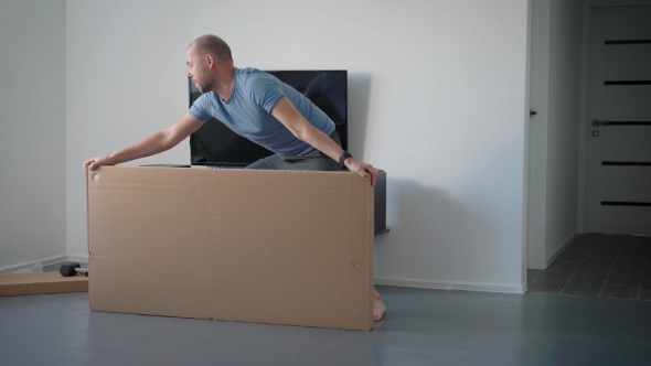 Adult Man Is Turning and Lying on a Floor Big Cardboard Box in an Room of Apartment, Cutting Packing alt