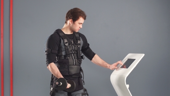 Man in Electrical Muscular Stimulation Suit Standing with Dumbbells Near Ems Tablet and Push on alt