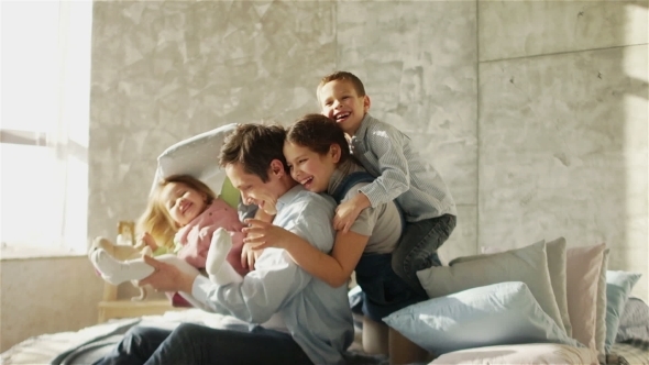 A Young Father and Three Children Embrace Sitting on the Bed. Happy Fatherhood. Father's Day alt