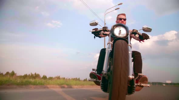 Stylish Biker with Tattoos Rides a Motorcycle on a Country Road at Sunset