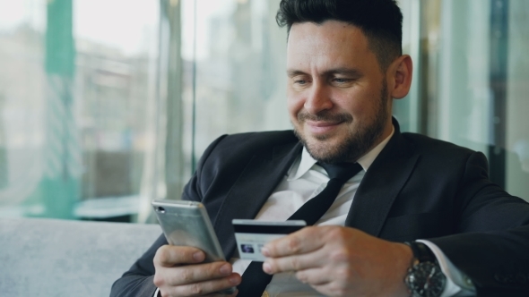 Happy Caucasian Businessman in Formal Clothes Paying Online Bill Keeping Credit Card and Smartphone alt