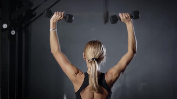 Sporty Young Blonde Woman Is Lifting Dumbbells By Hands in Gym, Standing Back To Camera alt