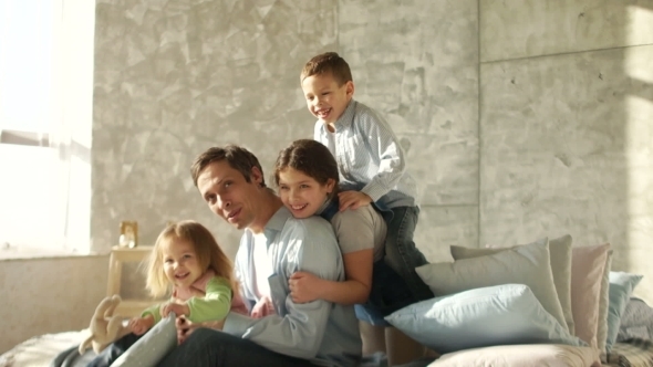 Three Children and a Young Father Embrace Sitting on the Bed. Family Morning. Happy Fatherhood alt