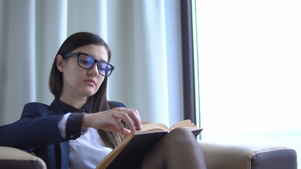 Young Attractive Business Woman in Office Suit Reads a Book Near the Window.