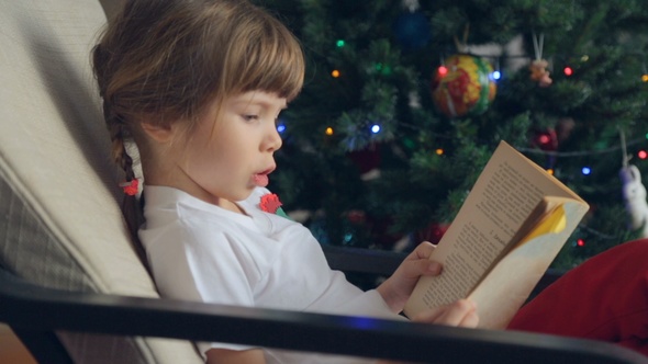 Small Girl Reading a Book in Front of Christmas Tree