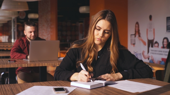 Business Young Girl Writing Note in Notebook in Cafe, Man with Laptop on Background