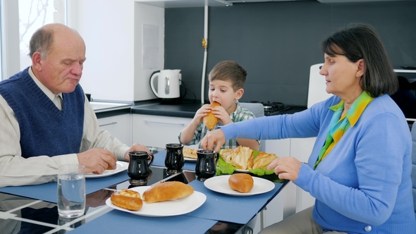 Pensioner, Child Eats Bakery Products with Grandfather and Grandmother in Kitchen alt