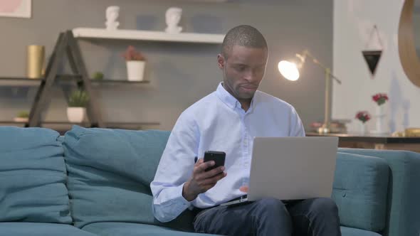 African Man Using Smartphone While Using Laptop on Sofa alt