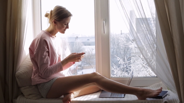 Young Beautiful Woman with a Smartphone on Window Sill. Winter Outside
