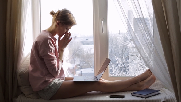 Sad and Upset Woman Using Her Laptop Sitting on Window Sill