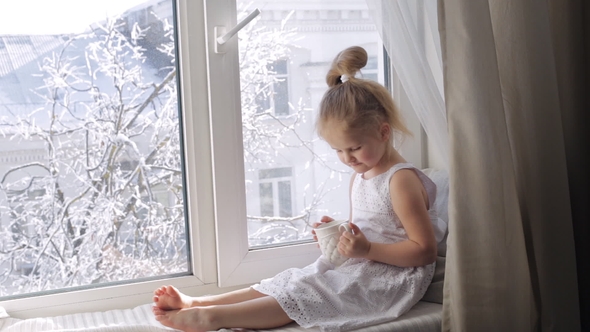 Cute Little Girl Drinks Tea Sitting on Window Sill. Sunny Winter Morning