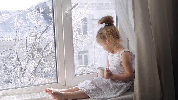 Cute Little Girl with a Cup of Tea Sitting on Window Sill. Girl with Cup Looking Through the Window