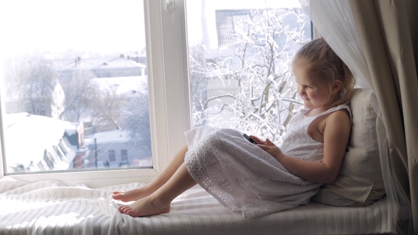 Funny Young Girl with Smartphone on a Window Sill. Sunny Winter Morning