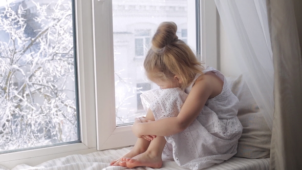 Sad Little Girl Looking Through the Window Sitting on Window Sill. Winter Outside