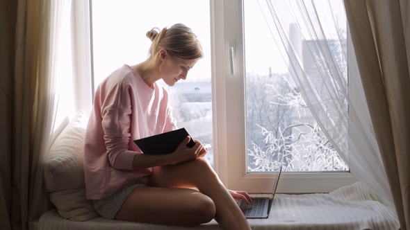 Young Woman with Notepad Working on Her Computer Sitting on Windowsill