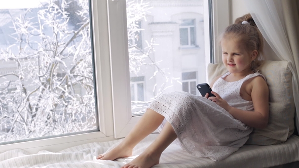 Young Girl Listens Music on Her Smartphone on a Window Sill. Sunny Winter Morning