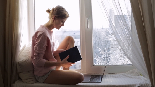Beautiful Young Woman with a Notepad Sitting on Window Sill. Winter Outside