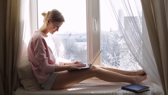 Woman Is Sitting on the Window Sill and Using Her Modern Laptop. Winter Outside the Window