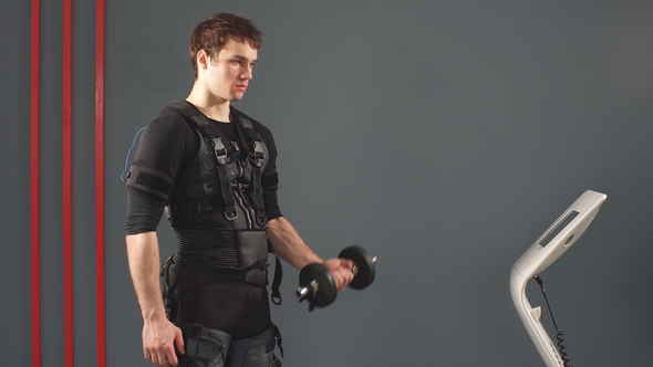 Fit Man in Electrical Muscular Stimulation Suit Standing with Dumbbells. alt