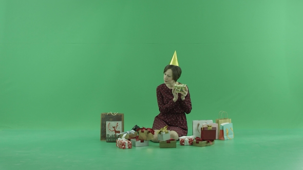 A Young Woman Is Sitting and Shaking Gift Boxes Around Her on the Green Screen