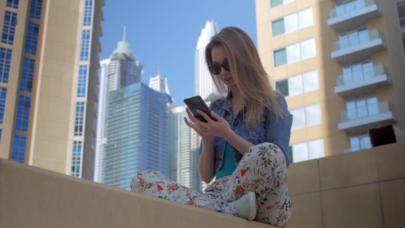 Teenage Girl Is Using Smartphone Sitting in Background of Modern Skyscrapers