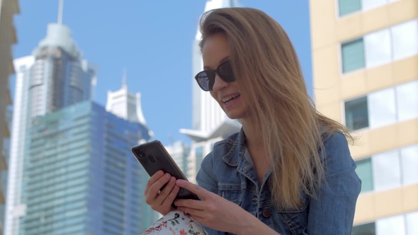 Smiling Blonde Girl Is Watching Funny Video on a Display of Her Phone, Sitting Outdoors