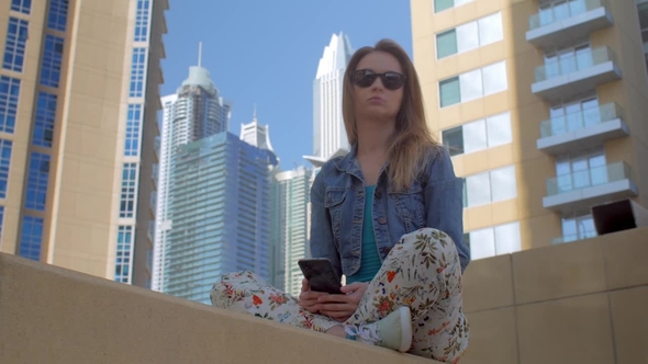 Young Woman with Long Blonde Hair Is Sitting in Background of Modern Buildings, Using Her Mobile