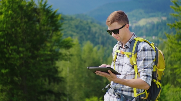 A Young Male Tourist with a Backpack Uses a Tablet. It Stands in a Picturesque Place Near the alt